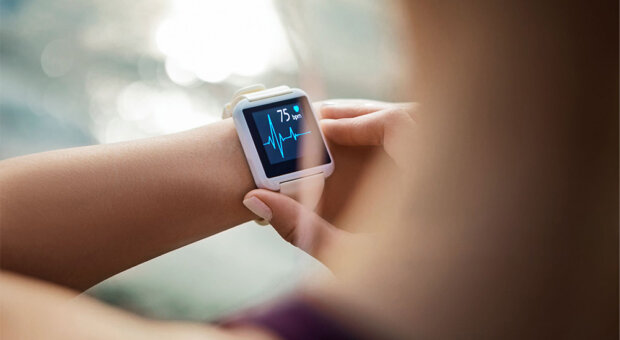 Close-up over the shoulder of a person checking a white smartwatch on their wrist, the screen showing a heart-rate reading of 75 bpm with a blue pulse line, outdoors in bright sunlight.
