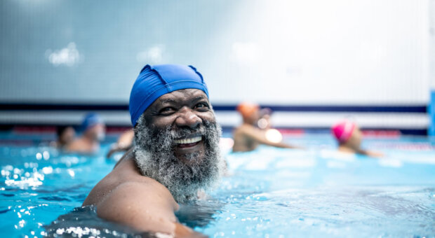 Older man with a full grey beard and a blue swim cap smiling at the camera while resting at the edge of an indoor pool, with other swimmers in coloured caps blurred in the lanes behind him.