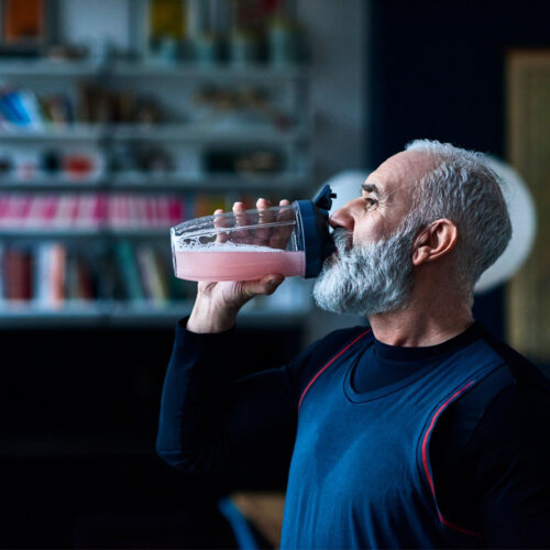Older man with grey hair and a full beard, wearing a navy athletic top, drinking a pink protein shake from a clear shaker bottle in a dimly lit room with bookshelves behind him.