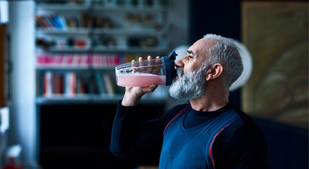 Older man with grey hair and a full beard, wearing a navy athletic top, drinking a pink protein shake from a clear shaker bottle in a dimly lit room with bookshelves behind him.